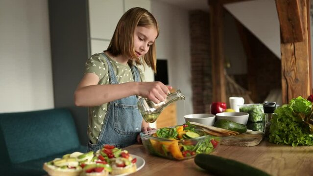 smiling little girl cooking a fresh healthy vegetable salad, pouring olive oil and mixing at the kitchen