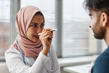 Medium close up of woman medical practitioner wearing hijab checking eyes of male patient with medical penlight