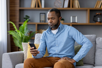Serious thinking mature man sitting on sofa at home, senior african american man holding phone,...
