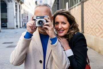 Mid adult tourist couple taking picture on vintage camera looking at camera. Senior woman hugging man while taking a photography in a city street