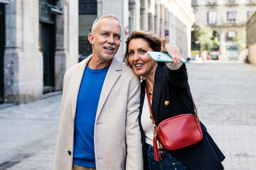 Carefree middle aged couple looking for touristic location pointing and holding smartphone. Happy...