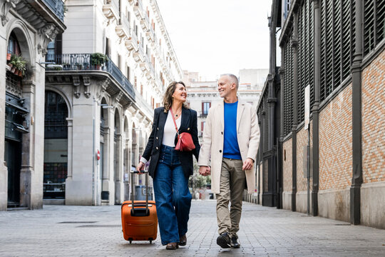 Affectionate mid adult tourist couple walking together with luggage in a european city. Lovely happy senior man and woman traveling with a suitcase visiting streets of old town.