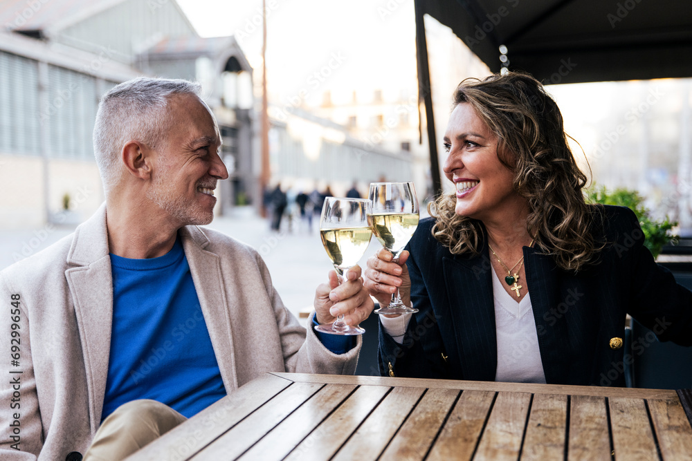 Wall mural Lovely mid adult couple toasting with wine while sitting in a terrace restaurant. Romantic date of a middle age woman and man drinking with glasses at a bar terrace looking each other.