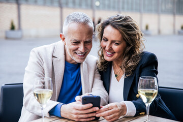 Joyful mature man and woman sharing smartphone while drinking wine in city at outdoors terrace bar. Cheerful handsome mid age couple using mobile phone together at a table restaurant.