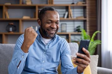 Online winner man sitting at home on sofa in living room, happy successful african american man...