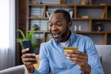 Joyful mature man at home on sofa with phone and bank credit debit card in hands, satisfied african...