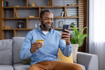 Joyful mature man at home on sofa with phone and bank credit debit card in hands, satisfied african...