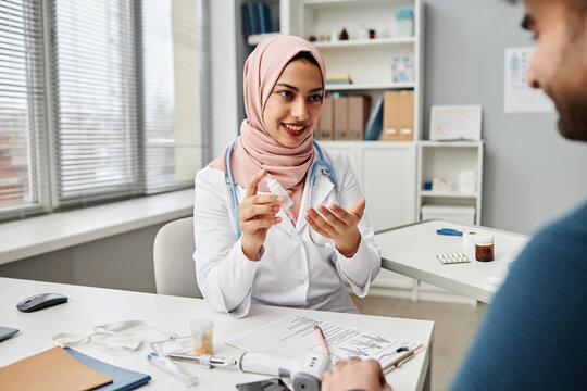 Portrait of young Muslim woman physician wearing pink headscarf advising eye drops to male patient during consultation in modern clinic office