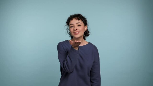 Joyful young curly woman sending air kiss to camera, flirting. Portrait smiling positive female in a blue sweater standing on isolated light blue background. People emotions lifestyle concept