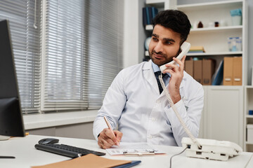 Medium shot of Muslim man doctor calling patient on landline phone while working at desk in modern clinic office