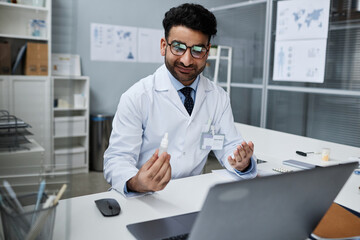 Medium shot of Muslim young male optometrist in glasses at desk in clinic office recommending eye drops to patient during online consultation - obrazy, fototapety, plakaty
