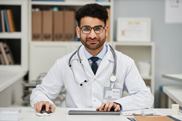 Portrait of young Islamic man physician in glasses looking at camera while working at desk using computer with hands on mouse and keyboard