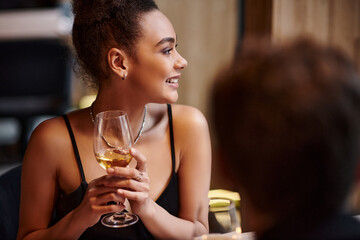 happy african american woman smiling and holding glass of wine during date on Valentines day