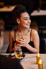 pleased african american woman smiling and holding glass of wine during date on Valentines day