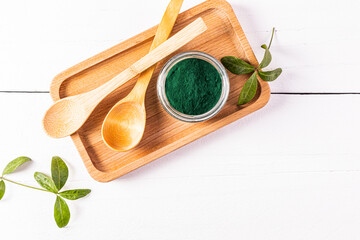 A glass jar filled with green spirulina algae powder and two wooden empty spoons on a wooden tray. Top view. White wooden table.