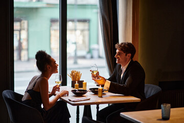 cheerful interracial couple in elegant attire holding glasses with wine during date in restaurant