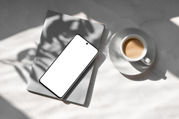 Mobile phone with blank screen mockup, gray notebook, cup with coffee drink and saucer on white...