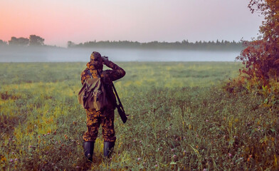 Man with gun stands on the edge of an autumn field in search of hunting object at dawn.