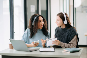 Two Attractive young female college students working on the school project using computer and tablet together, enjoy talking and headphones having a video chat.