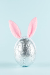 Easter egg wrapped in a silver foil with pink bunny ears against pastel blue background. Easter minimal concept. Creative Happy Easter or spring layout.