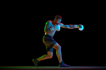 Young muscular man, shirtless boxing athlete in motion, training, practicing hooks isolated over black background in neon light. Concept of professional sport, combat sport, martial arts, strength