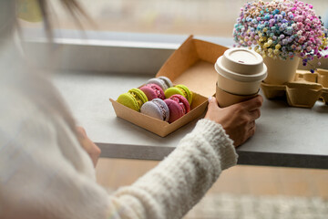 Female hand holds a cardboard cup with tea next to a box of macaroons