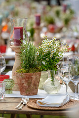 Dining table setting with vase of flowers, bowls, glasses, candles, cutlery, with blurred background.