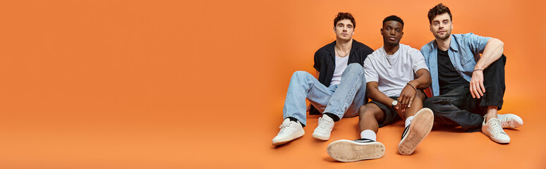 handsome multiracial stylish men in trendy urban outfits sitting on floor, fashion concept, banner