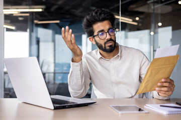 Worried young Indian businessman man sitting in the office at the table and reading the received letter and documents, frustrated and confused throwing up his hands