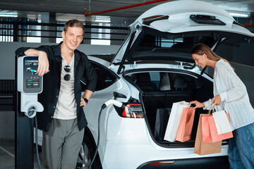 Young woman travel with EV electric car to shopping center parking lot charging in downtown city...