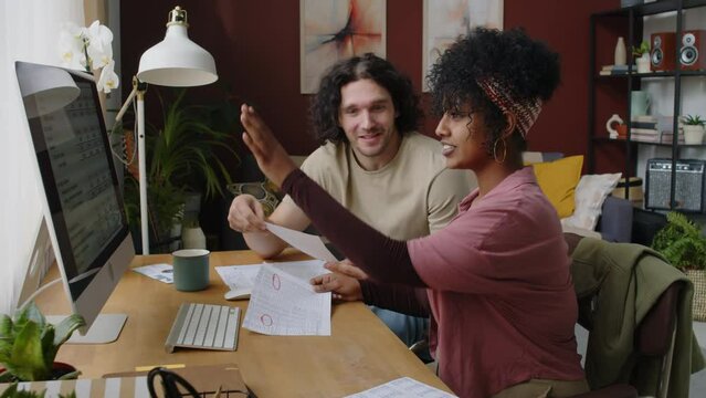 Medium shot of young interracial couple sitting at table by desktop computer and discussing total amount of their spends, reading and counting paper receipts, checks and bills