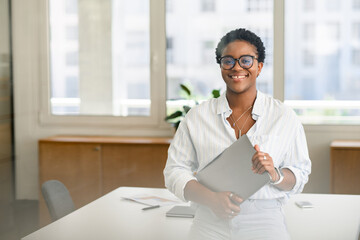 Carefree inspired african-american female employee standing in modern office space and holding...