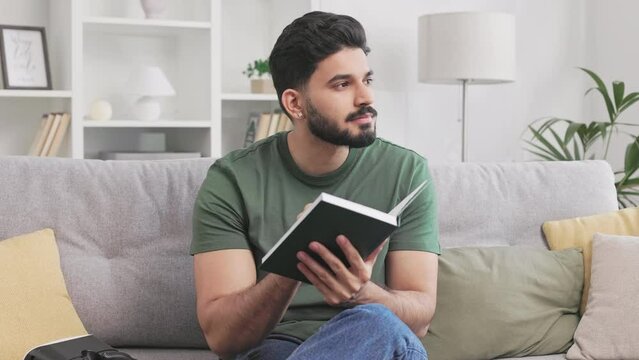 Handsome bearded guy using personal copybook for noting thoughts while sitting on couch and looking pensively aside. Indian man in casual attire enjoying free time alone at home with new insights.