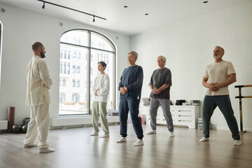 Wide angle shot of senior people standing in gym and doing qigong exercise for energy concentration while male master guiding them - obrazy, fototapety, plakaty