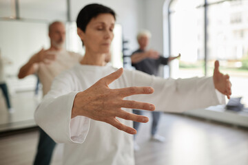 Medium shot with focus on hand of senior female student doing qigong concentration exercise in gym - obrazy, fototapety, plakaty