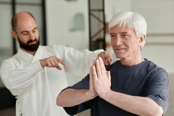 Medium shot of white-haired senior man doing prayer pose exercise while qigong master observing pose accuracy