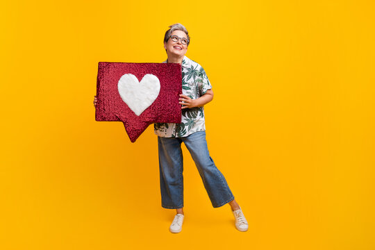Full length photo of lovely grandma hold heart icon look empty space wear trendy tropical print garment isolated on yellow color background