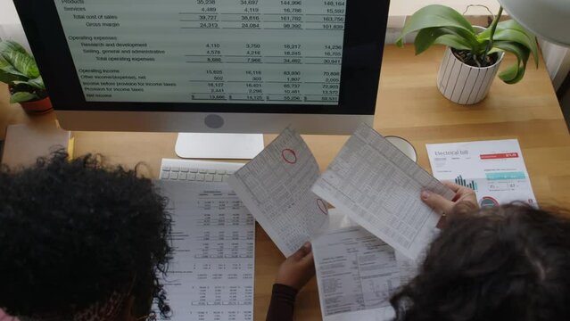 Top view of young couple looking through piles of financial documents, bills and receipts while managing and planning household budget together sitting at table by desktop computer