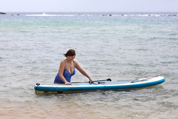 Back view of adult overweight woman in swimsuit with SUP board and paddle goes into sea. Wellness middle aged female enjoys surfing on paddle board on summer vacation. Active modern lifestyle.