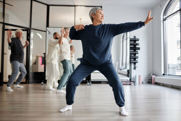 Full shot of gray-haired senior woman dressed in blue sweatsuit practicing qigong archer pose in...