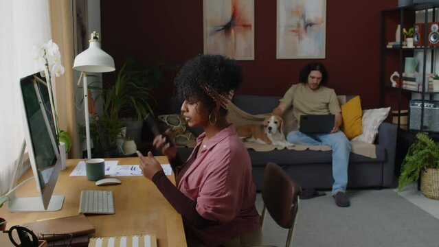 Medium shot of young Biracial businesswoman using desktop computer and making phone calls while working remotely from home office with her Caucasian husband and their dog sitting on sofa in background