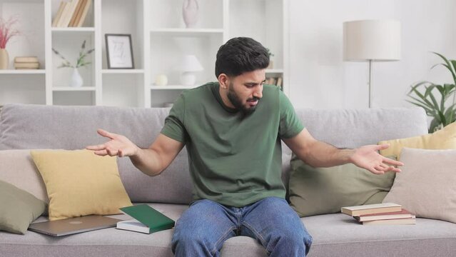 Sad hindu guy dressed in casual clothes sitting on couch among scattered books and shrugging shoulders while saying I don't know. Concept of stress, education and people.