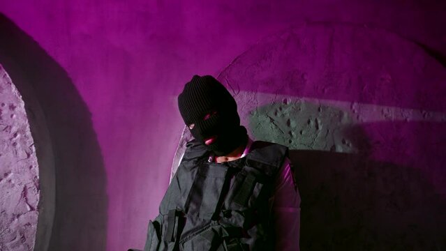 woman gangster or soldier in balaclava and bulletproof vest standing alone near wall, portrait