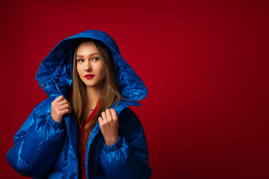  Fashion portrait of beautiful confident woman wearing trendy winter oversized blue puffer down jacket with hood, posing in studio on red background. Copy, empty space for text