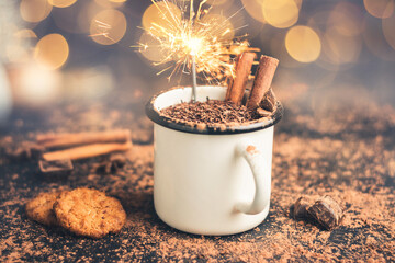 Homemade hot chocolate drink with cinnamon stick, star anise, grated chocolate and sparklers in enamel mug on dark background with cookies, cacao powder and chocolate pieces, Christmas lights bokeh - obrazy, fototapety, plakaty