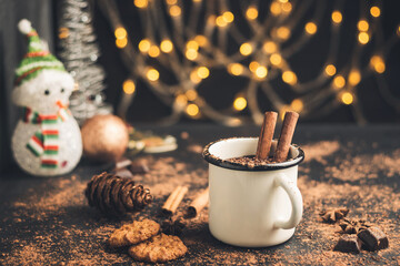 Homemade spicy hot chocolate drink with cinnamon stick, star anise, grated chocolate in enamel mug on dark background with cookies, cacao powder and chocolate pieces, Christmas lights bokeh - obrazy, fototapety, plakaty