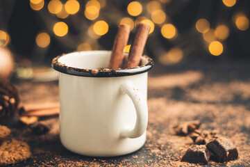 Homemade spicy hot chocolate drink with cinnamon stick, star anise, grated chocolate in enamel mug on dark background with cookies, cacao powder and chocolate pieces, Christmas lights bokeh