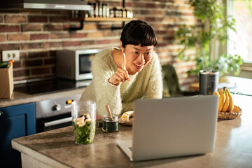 Asian Woman Preparing a Green Smoothie while Video Calling on Laptop in a Modern Kitchen