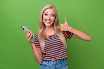 Photo of adorable optimistic girl wearing trendy clothes thumb up nice offer price gadget isolated on green color background