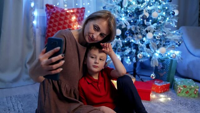 Mother and son, smiling, take pictures on the phone under the Christmas tree. Lights shine, balls and toys create a magical atmosphere in which every moment is a festive fairy tale. 